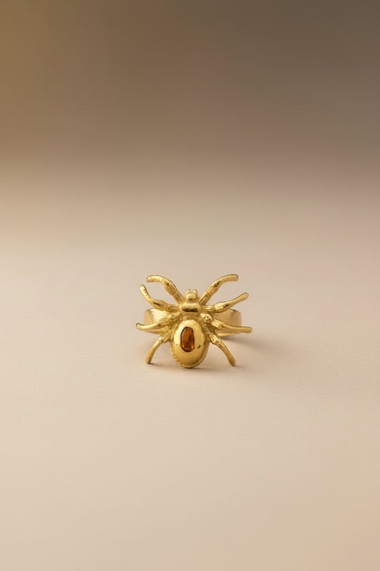 Gold spider-shaped ring with a red gemstone on a light gray background
